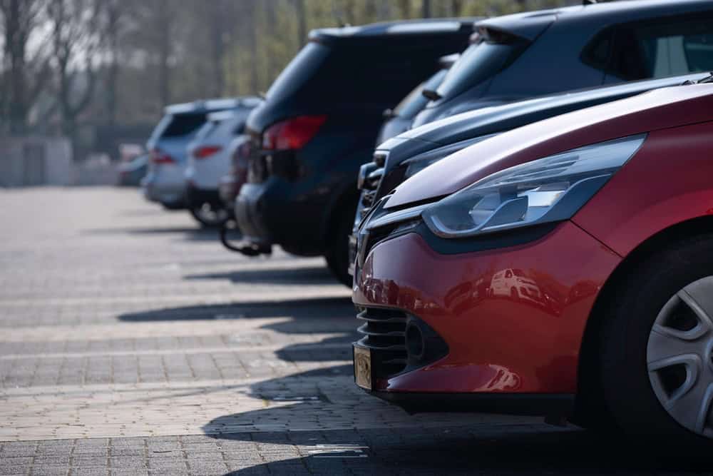 A row of parked vehicles in an outdoor parking lot under daylight, with the front end of a red car prominently in the foreground. Other cars of various colors are lined up beside it. Sunlight filters through nearby trees, casting soft shadows across the pavement. The scene suggests recently serviced vehicles at a trusted auto repair center in Clarke County.