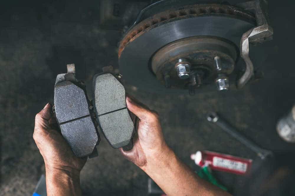 Two hands hold brake pads side by side in front of a vehicle’s brake disc. The left brake pad appears new with thick friction material, while the right pad is worn and noticeably thinner. The background shows part of the wheel hub and a tube of grease, highlighting detailed brake maintenance in Clarke County.