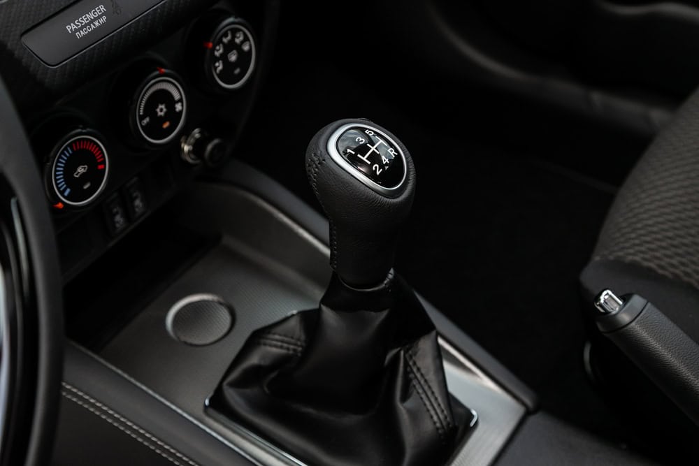 Close-up of a car’s manual gear shifter with a black interior. The shift knob clearly shows the gear positions engraved on its top, and the black leather shift boot extends below it. Surrounding the gear shifter are climate control dials and part of the center console, including a cup holder, illustrating the vehicle’s functional interior during an auto repair inspection in Clarke County.