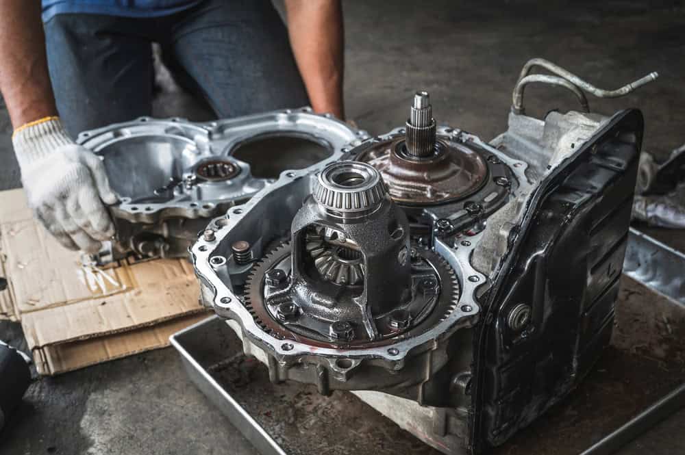 A mechanic wearing protective gloves works on a disassembled car gearbox, handling the gearbox housing. Various internal components, including gears and shafts, are neatly arranged on a metal workbench and a piece of cardboard nearby. The scene depicts careful inspection and repair of automotive transmission parts in a professional Clarke County workshop.