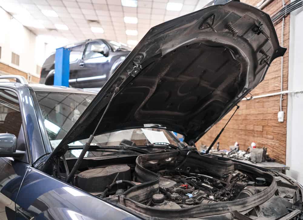 A car with its hood open is positioned in the foreground of a brightly lit auto repair shop. In the background, another vehicle is elevated on a red hydraulic lift, likely undergoing brake repair services in Clarke County. The garage is equipped with toolboxes, diagnostic machines, and maintenance equipment, giving the space a professional and organized look.