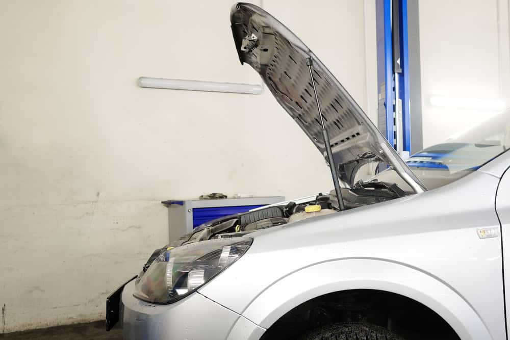 A silver car with its hood open is parked inside a garage, likely a site for Auto Repair Clarke County. A blue and white toolbox is visible in the background against a white wall. The car's front right headlight and part of its engine are visible, indicating it might be undergoing maintenance or repair work.