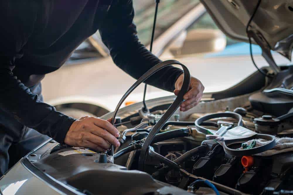 A person wearing a dark shirt works on a car engine with the hood open, holding a serpentine belt stretched between their hands. Various engine parts and tools surround the workspace, highlighting the careful handling involved in installing or removing the belt during auto repair in Clarke County.