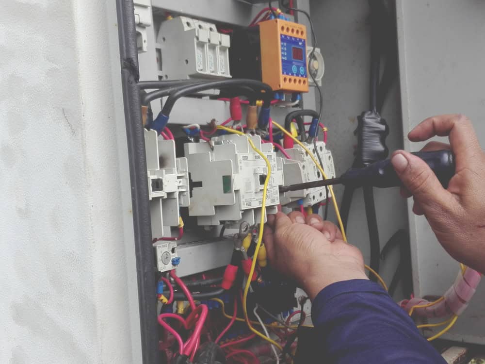 A close-up of a person's hands using a screwdriver to adjust or secure wiring inside a detailed electrical panel. The panel is filled with an array of multicolored wires—red, blue, yellow, and black—connected to various terminals, switches, and circuit components. The person is focused on a specific section, indicating precision work commonly associated with transmission diagnostics or electrical system repair in Clarke County.