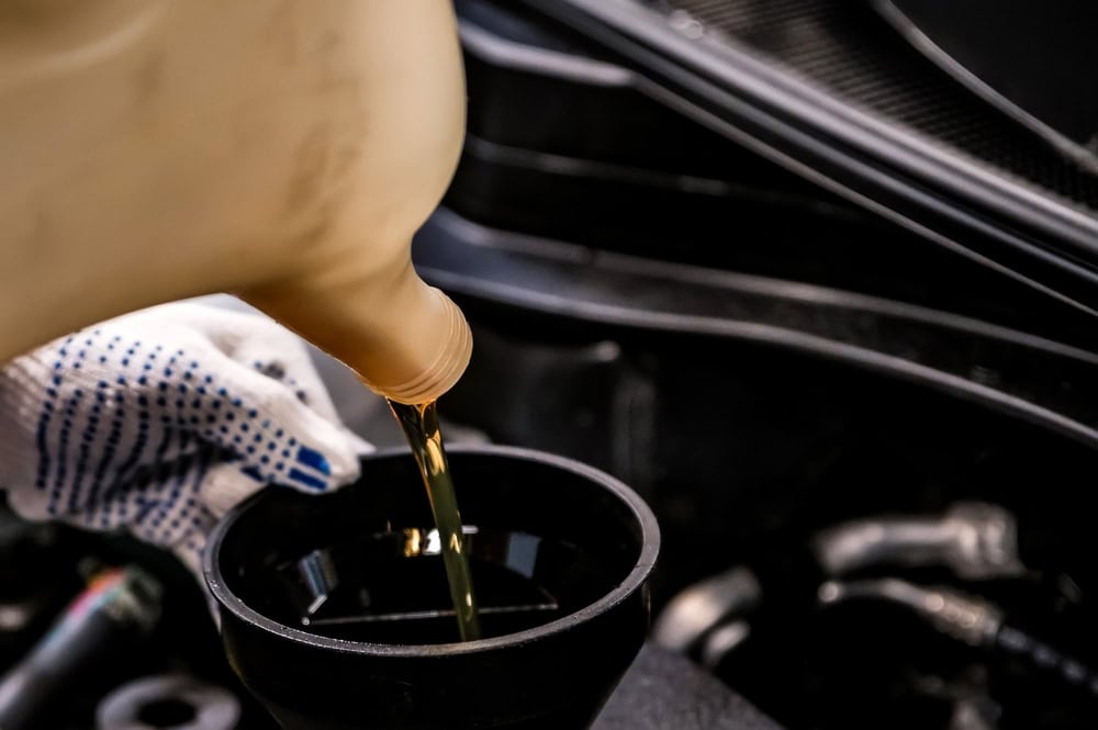 A person wearing a white glove with blue rubberized dots is carefully pouring golden motor oil from a translucent plastic container into a black funnel placed inside a car’s engine compartment. The funnel sits securely in the oil fill port, and surrounding engine components are visible, illustrating the precision and cleanliness involved in a professional oil change service in Clarke County.