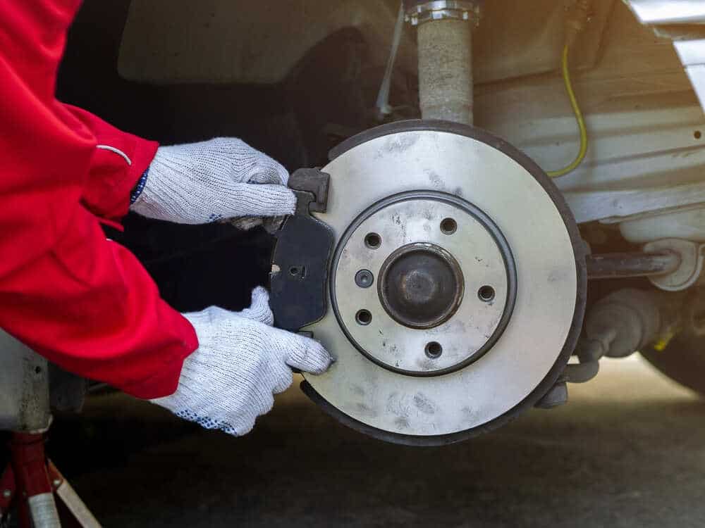 A mechanic wearing white gloves and a red jacket installs or inspects a brake pad on a car’s disc brake rotor. The close-up view captures the mechanic’s gloved hands working on the brake assembly, with the rotor, caliper, and surrounding components clearly visible demonstrating expert brake repair in Clarke County.