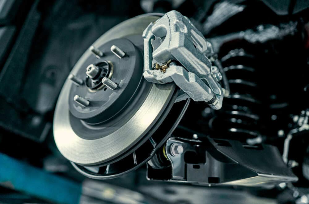 Close-up view of a car’s brake system serviced by Auto Repair Clarke County, showing a clean, shiny ventilated disc brake paired with a silver caliper. Suspension components are slightly visible in the background under bright lighting that highlights the detailed metalwork.