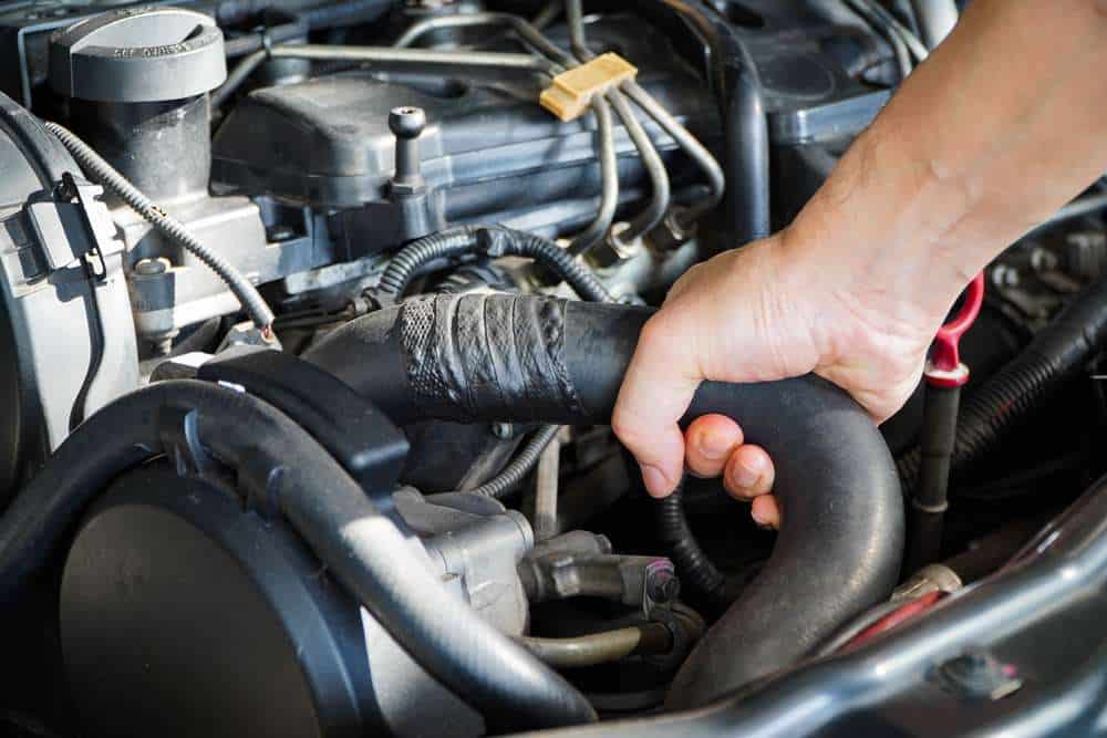 A close-up of a hand gripping a black rubber hose inside a car’s engine bay. The hose is wrapped and secured with tape, surrounded by various wires, metal components, and engine parts, highlighting a moment of focused car maintenance or engine repair in Clarke County.