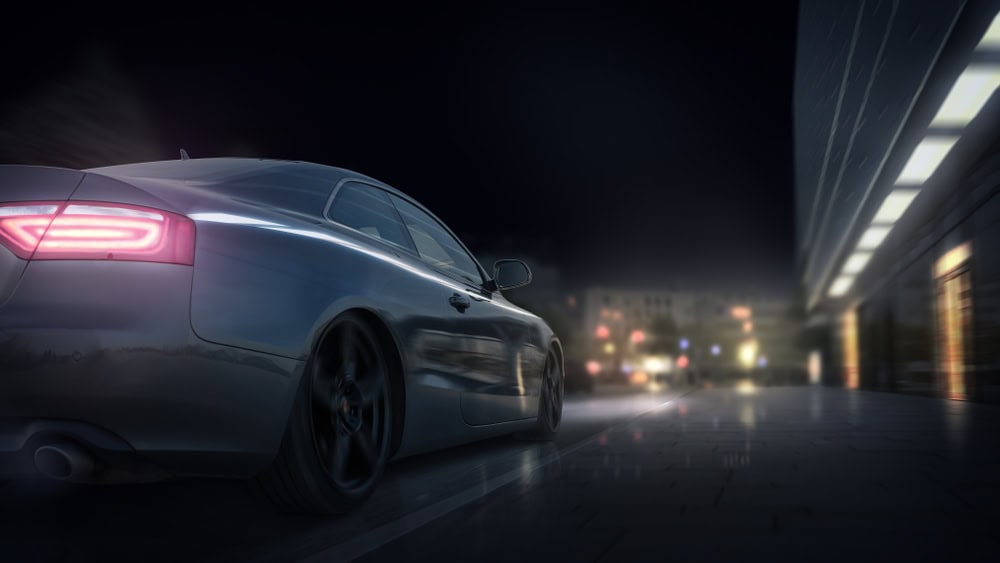 A sleek silver sports car with glowing red taillights speeds down a rain-slicked city street at night. Reflections from neon signs, streetlights, and modern glass buildings shimmer across the wet pavement, creating a dramatic, futuristic atmosphere. The car is in motion, captured from the rear as it drives away, possibly en route to an auto repair shop in Clarke County.