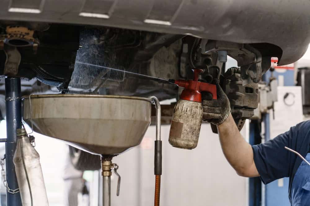A mechanic in a well-lit auto repair shop uses a spray bottle to clean or treat parts beneath a car that’s lifted on a hydraulic lift. Spilled automotive fluid is being collected in a wide, industrial funnel connected to a container below the vehicle. The workspace includes scattered tools, hoses, and repair equipment, reflecting a typical scene from a professional auto repair service in Clarke County.