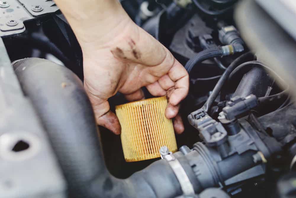 Close-up of a mechanic's dirty hand holding a used yellow engine oil filter over an open car engine bay. The filter is stained with dark oil, indicating it has been removed for replacement. Surrounding engine components like hoses, clamps, and the filter mount are visible, showing that oil maintenance is actively underway at an auto repair shop in Clarke County.