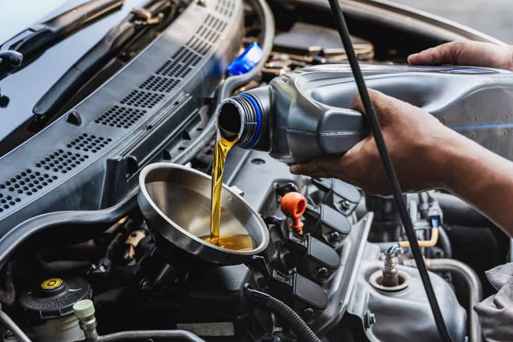 A person is pouring motor oil into a car engine using a funnel. The car's hood is open, revealing the engine components. The individual appears to be performing routine maintenance or an oil change on the vehicle, perhaps following advice from a Mechanic Clarke County specialist for optimal engine care.