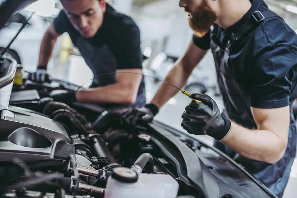 Two mechanics in dark clothing and gloves are working under the hood of a car. One is inspecting the engine while the other holds a dipstick to check the oil level. The setting appears to be a well-lit auto repair shop in Clarke County, specializing in thorough engine repairs and oil changes.