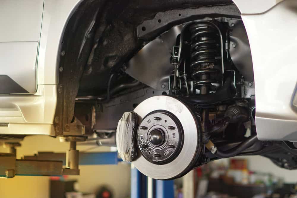 Close-up view of a car's front suspension and braking system at Mechanic Clarke County. The car is elevated on a hydraulic lift, revealing the exposed coil spring, shock absorber, and brake disc. The wheel has been removed, giving a clear look at the mechanical components.