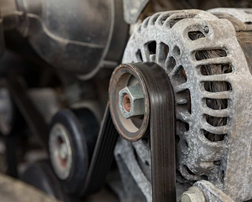 Close-up of a car alternator with a serpentine belt wrapped around it, showcasing typical wear and tear seen in Auto Repair Clarke County. The surface of the alternator has a layer of dust and dirt, indicating use. Other engine components can be seen in the background, slightly out of focus.
