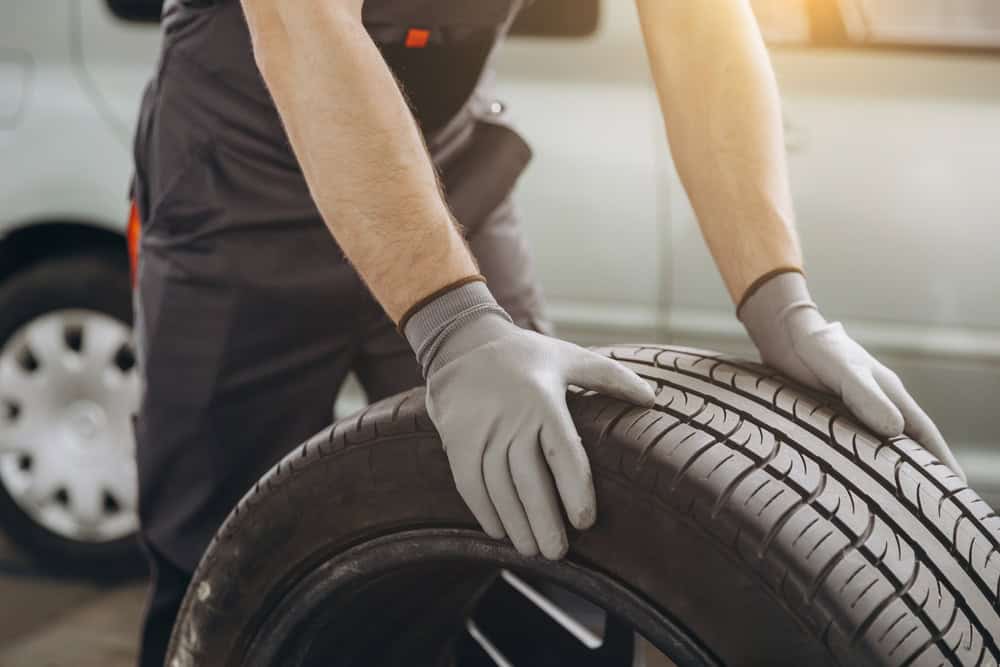 A person wearing gloves is handling a car tire in what appears to be an automotive workshop. A silver car with one of its wheels off is in the background, capturing a mechanic's tire replacement or maintenance. This scene could easily be part of services like engine repair Clarke County.