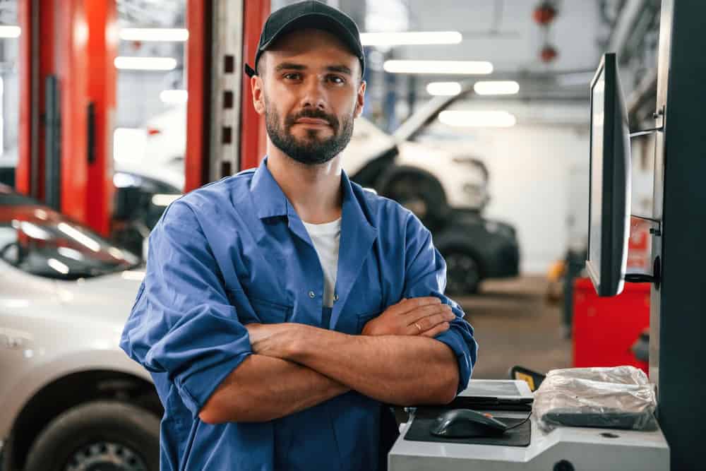 A mechanic in a blue jumpsuit and black cap stands with folded arms in an auto repair shop. In the background, there are vehicles and repair equipment, including a car lifted on a hydraulic lift. Specializing in transmission repair Clarke County residents trust, he's ready for any challenge.