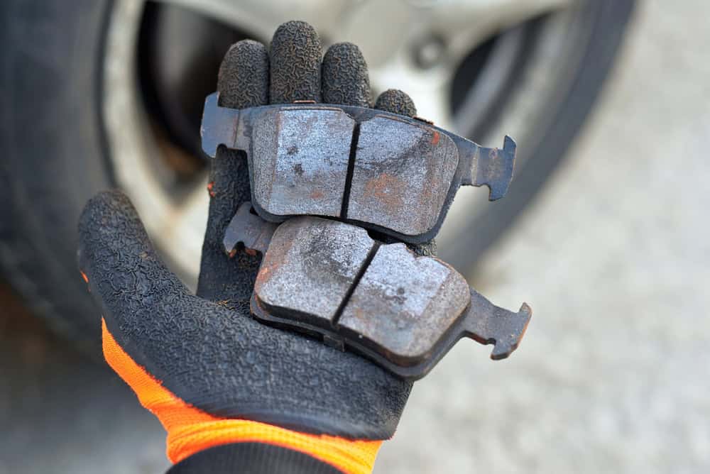 A close-up of a gloved hand—wearing a black and orange mechanic’s glove—holding two heavily worn and rusted brake pads in front of a car wheel. The pads show deep grooves, corrosion, and thinning edges, clearly indicating they are due for replacement. The background includes the vehicle’s wheel hub and part of the tire, slightly blurred to emphasize the brake pads. The glove’s professional design suggests the hand belongs to an experienced mechanic, likely from a trusted Clarke County repair shop.