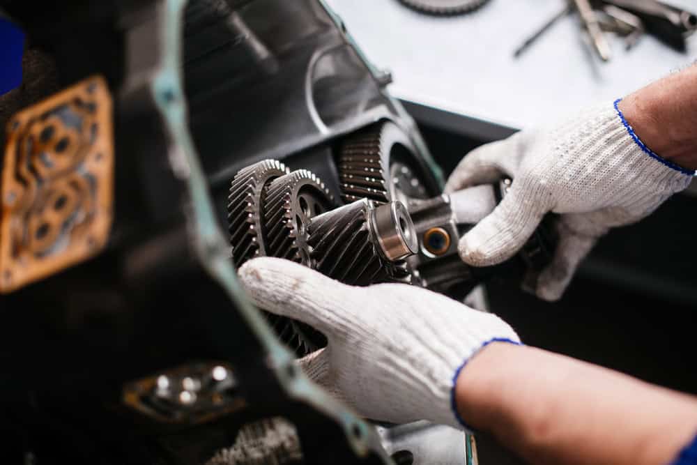Close-up of a person's hands wearing white gloves while assembling or repairing a set of metallic gears in a mechanical device, embodying the meticulous attention to detail seen in auto repair in Clarke County. Other tools and gear parts are visible in the blurred background.
