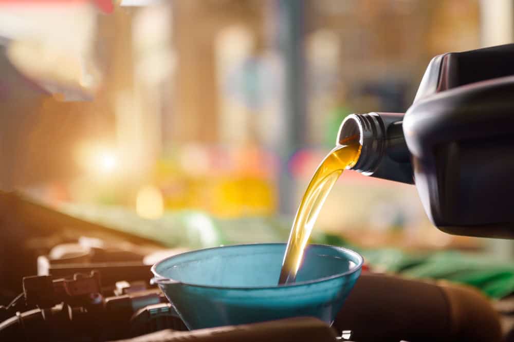 A close-up of a blue funnel being used to pour engine oil from a black container. The oil is a golden color, and the background is blurred with warm light coming from the left, suggesting it might be taken in a brightly lit garage or workshop in Clarke County during an auto repair.