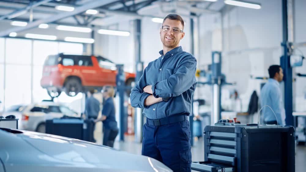 A confident mechanic wearing safety glasses and a blue uniform stands with arms crossed inside a busy auto repair shop in Clarke County. Behind him, a red SUV is raised on a hydraulic lift for an oil change while other technicians work. The scene includes tools, equipment, and workbenches, emphasizing a professional garage environment.