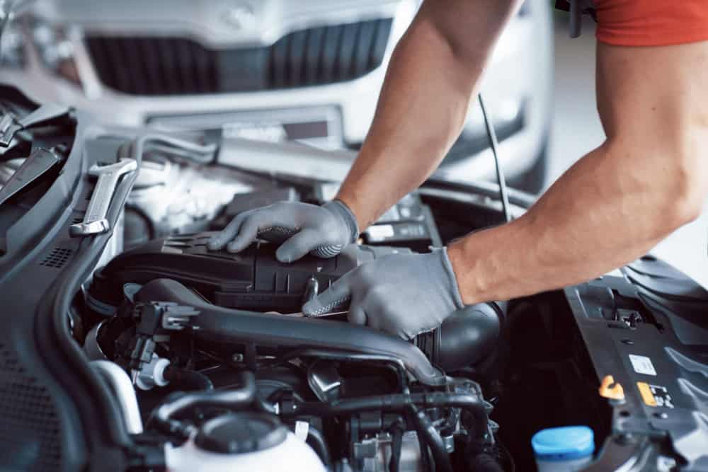 A close-up of a mechanic's gloved hands working on an engine under the hood of a car in Clarke County. The focus is on the engine components as the mechanic uses tools to make adjustments, perhaps during an oil change or transmission repair. A blurred vehicle is visible in the background.