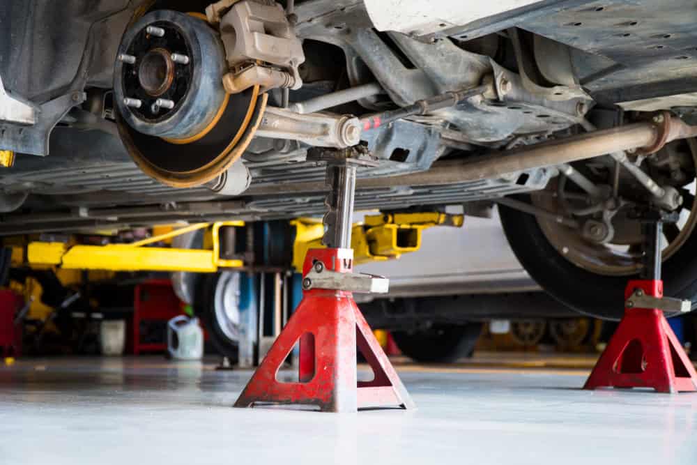 A vehicle is elevated on red jack stands in a garage, revealing its undercarriage. The brake assembly and suspension components are visible, and the car appears to be undergoing engine repair in Clarke County. Various tools and equipment are seen in the background.