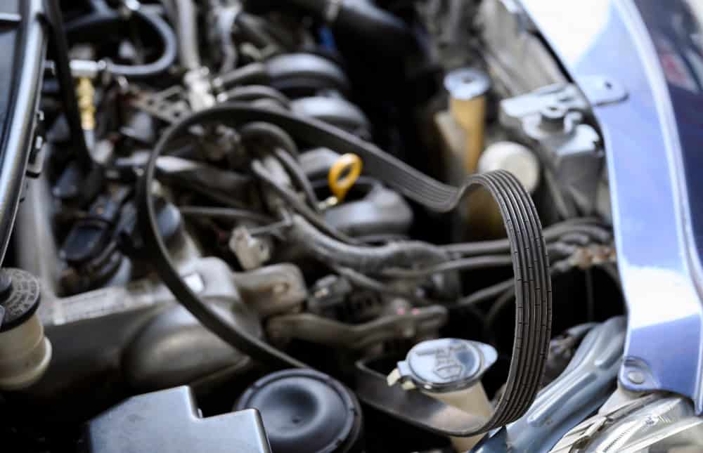 Close-up of a car engine with various components visible, including a large, visible belt in the foreground. The engine bay shows cables, hoses, and metal parts, along with the oil dipstick and other engine parts, indicating a complex mechanical system—ideal for an Oil Change Clarke County service.