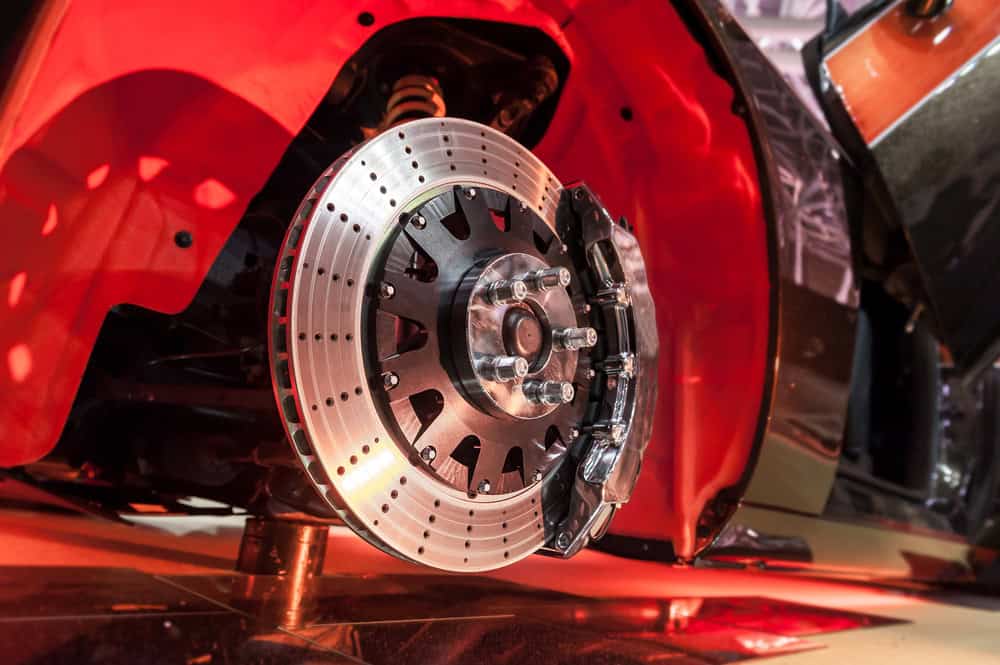 A close-up of a high-performance car’s exposed brake system. The image features a large, drilled and slotted brake rotor paired with a performance-grade caliper. The red car body contrasts with visible black suspension and chassis components, showcasing the precision and complexity of modern automotive engineering.