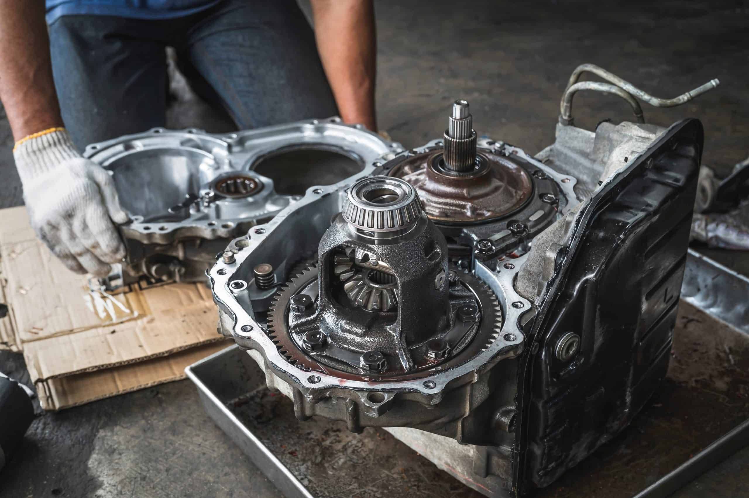 A person wearing gloves is working on a car part, possibly the transmission, in what seems to be a Transmission Repair Clarke County workshop. Various mechanical components are visible, with the person holding a part and tools scattered around on a cardboard surface. The setting appears to be a dedicated mechanic's garage.