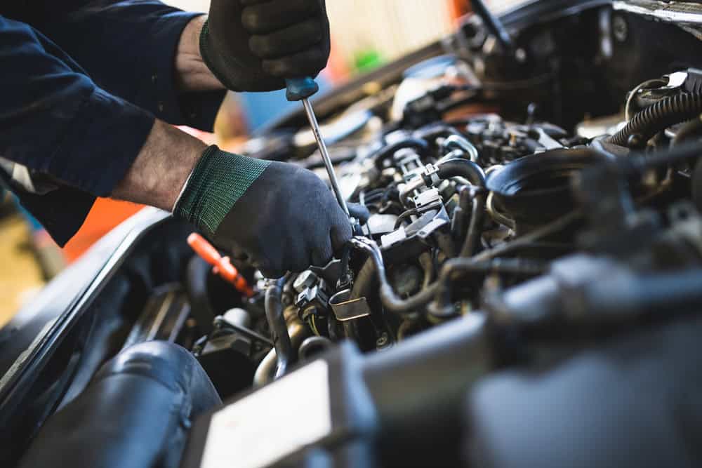 A person wearing black gloves is working on a car engine with a screwdriver. The close-up shot focuses on the hands and engine components, depicting a mechanic in Clarke County expertly repairing or maintaining the engine.