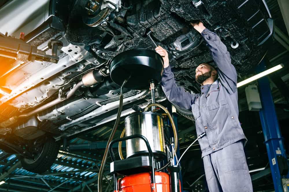 A mechanic in a gray jumpsuit works beneath a lifted vehicle inside a garage. They use a tool to perform maintenance on the car’s underside. Around the work area, various tools and equipment are visible, reflecting a well-equipped and organized auto repair shop in Clarke County.
