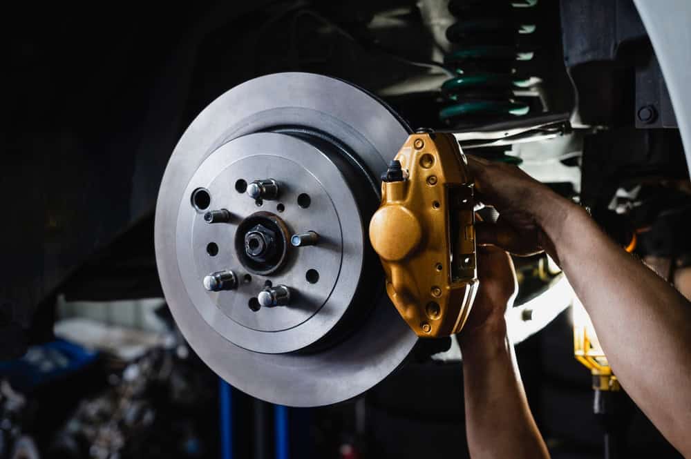 A close-up of a car’s brake system undergoing repair in Clarke County. A person’s hands are working on a shiny silver brake disc and a vibrant yellow caliper. Visible in the background are components of the suspension system and various tools, emphasizing the precision and skill involved in professional auto repair.