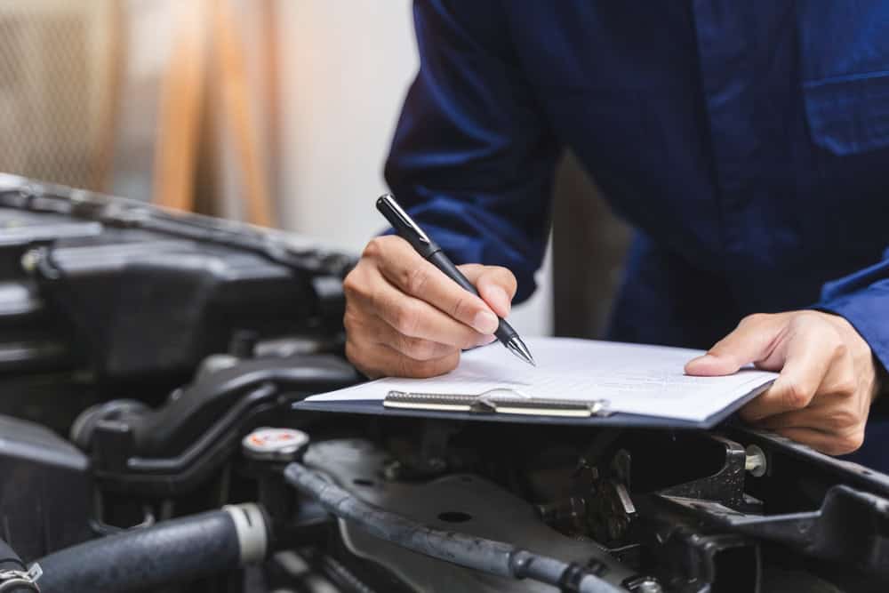 A person wearing a blue uniform writes on a clipboard resting on a car engine. The image highlights their hand holding a pen, with various engine components and car parts visible around the workspace—capturing a moment of documentation during engine repair in Clarke County.