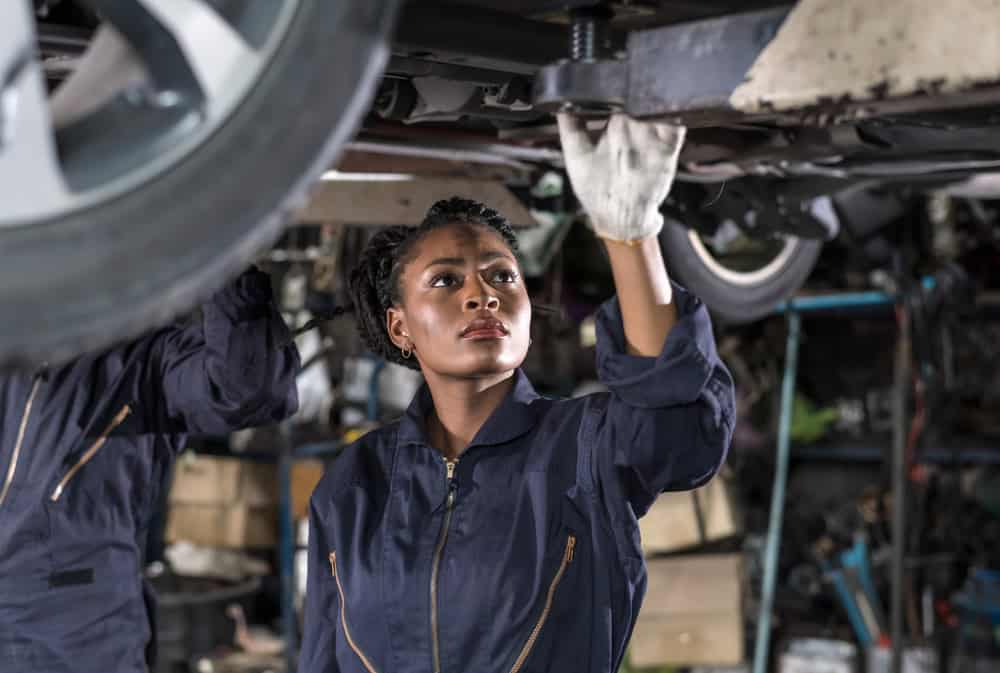 A mechanic wearing a dark blue coverall and white gloves carefully inspects the underside of a vehicle raised on a lift inside a professional auto repair shop. His focused expression and steady posture convey precision and attention to detail. The garage setting, located in Clarke County, includes visible shop tools and equipment in the background, reinforcing a hands-on, expert service environment.