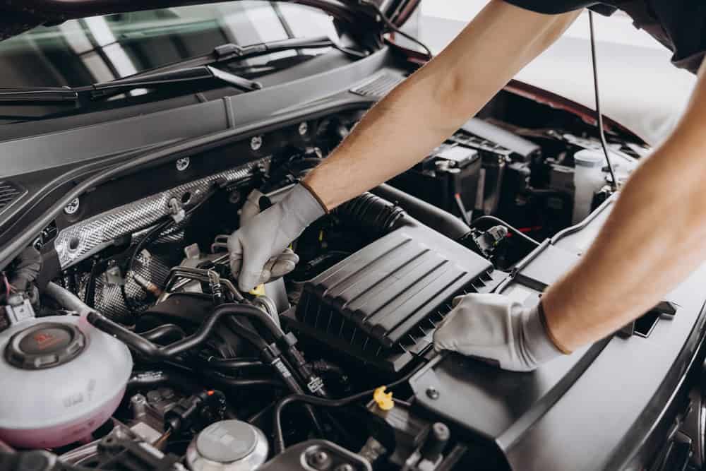 A person wearing gloves uses tools to adjust components inside an open car engine. The focus is on their hands and the engine bay, with visible hoses, belts, and mechanical parts. The scene captures a moment of skilled engine repair work taking place in a professional auto repair shop in Clarke County.