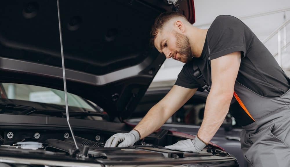 A bearded mechanic wearing a black t-shirt and grey overalls works under the raised hood of a car in an auto repair shop, possibly in Clarke County. His hands are actively engaged in inspecting or repairing components within the engine bay. The scene captures his focus and hands-on approach, reflecting the thorough and skilled service typical of a professional automotive workshop.