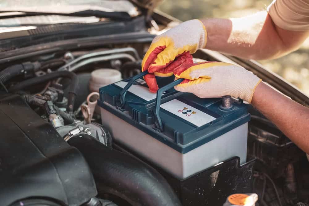 A person wearing yellow and red gloves lifts a car battery using a red cloth from the engine bay of a vehicle with the hood open. Various engine components surround the battery area, indicating an ongoing service. The scene takes place outdoors, showcasing hands-on automotive expertise provided by Auto Repair Clarke County.