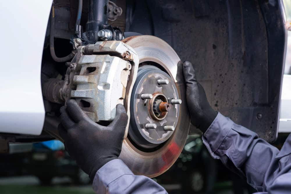 A mechanic wearing black gloves services the front disc brake system of a car raised on a lift in a professional garage. With the wheel removed, the brake rotor and caliper are clearly visible. The mechanic’s hands are positioned on the rotor, indicating precise adjustment or inspection. The setting reflects the clean, organized environment of a trusted auto repair facility in Clarke County.