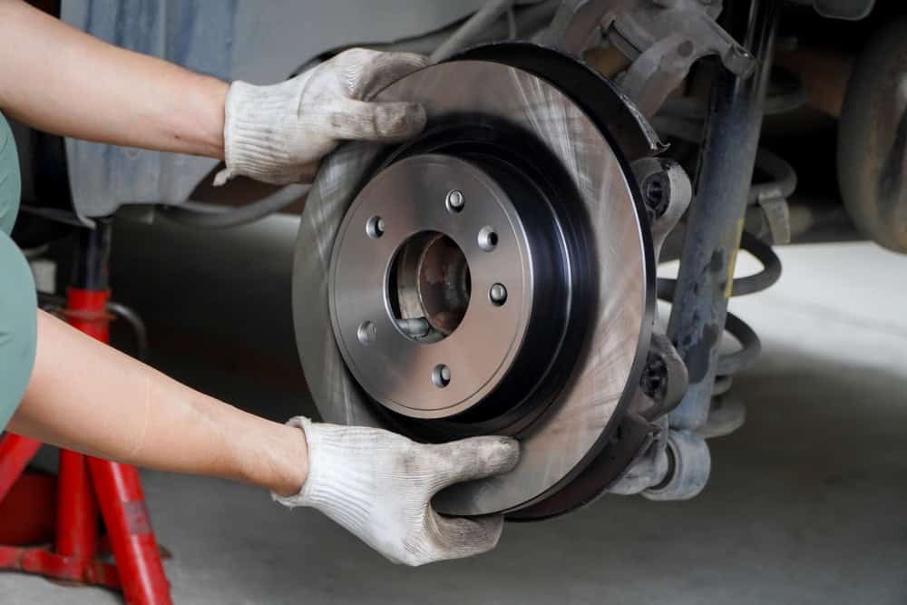 A mechanic wearing gloves installs a shiny new brake disc on a vehicle elevated on a red jack. The car is lifted in a professional garage setting—Auto Repair Clarke County—where the brake caliper, suspension components, and part of the wheel hub are clearly visible in the background. The scene emphasizes precision, safety, and expert-level brake system service.