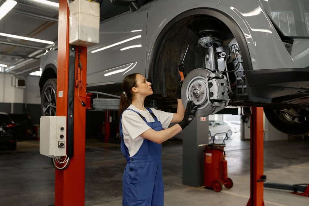 A female mechanic wearing blue overalls works on the brake system of a car lifted on a red hydraulic lift inside an auto repair shop. She uses a wrench with focused precision, embodying the hands-on expertise of Brake Repair Clarke County. Other vehicles, tools, and equipment are visible in the background, reinforcing the busy and professional environment.
