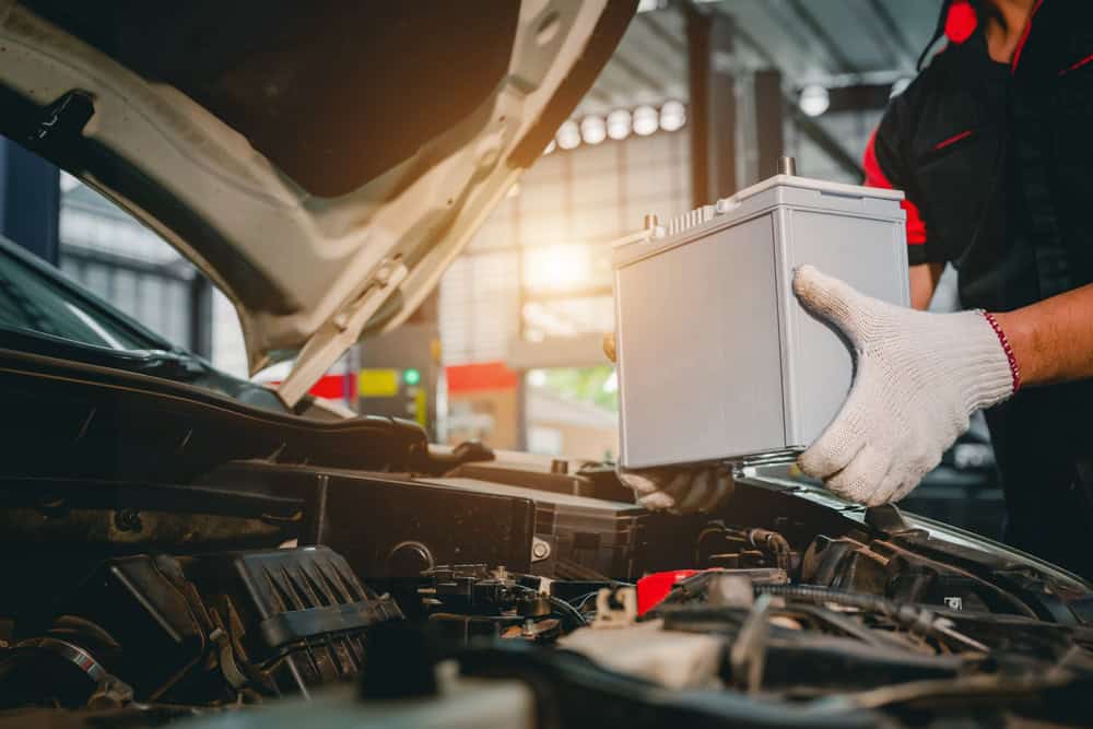 A Mechanic Clarke County-based technician wearing white gloves installs a car battery under the open hood of a vehicle in a workshop. Sunlight filters through the windows, illuminating the intricate components of the engine compartment.