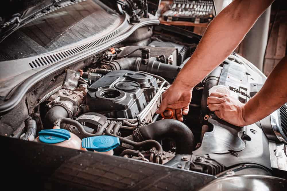 A mechanic in dark workwear leans over the open hood of a car, examining the engine with both hands engaged on various components. The engine bay features visible blue caps and metallic engine parts, indicating active diagnostics or maintenance work. In the softly lit background, workshop tools and equipment are faintly visible, reinforcing the setting as a professional automotive repair facility. The mechanic’s focused posture and hand placement suggest deep technical expertise and attention to mechanical precision.