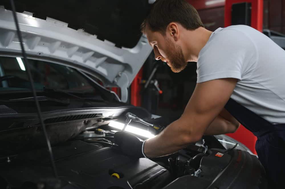A mechanic wearing gloves and a white t-shirt holds a wrench while working on a car engine under the open hood inside a garage. A bright work light casts focused illumination on the area, highlighting his concentrated expression and steady hands as he performs repairs. The surrounding setting includes shadows, tools, and the structured interior of a garage—emphasizing the hands-on, detail-oriented nature of professional auto service at Pro Transmissions & Automotive of Athens.