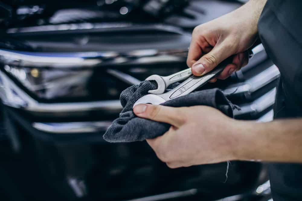 A close-up view of a person's hands meticulously cleaning an automotive tool with a dark cloth. The hands show focused care and precision, suggesting routine maintenance or preparation for repair. In the softly blurred background, part of a car is visible, subtly indicating a workshop or garage setting. The composition emphasizes attention to detail and pride in craftsmanship hallmarks of service at Pro Transmissions & Automotive of Athens.