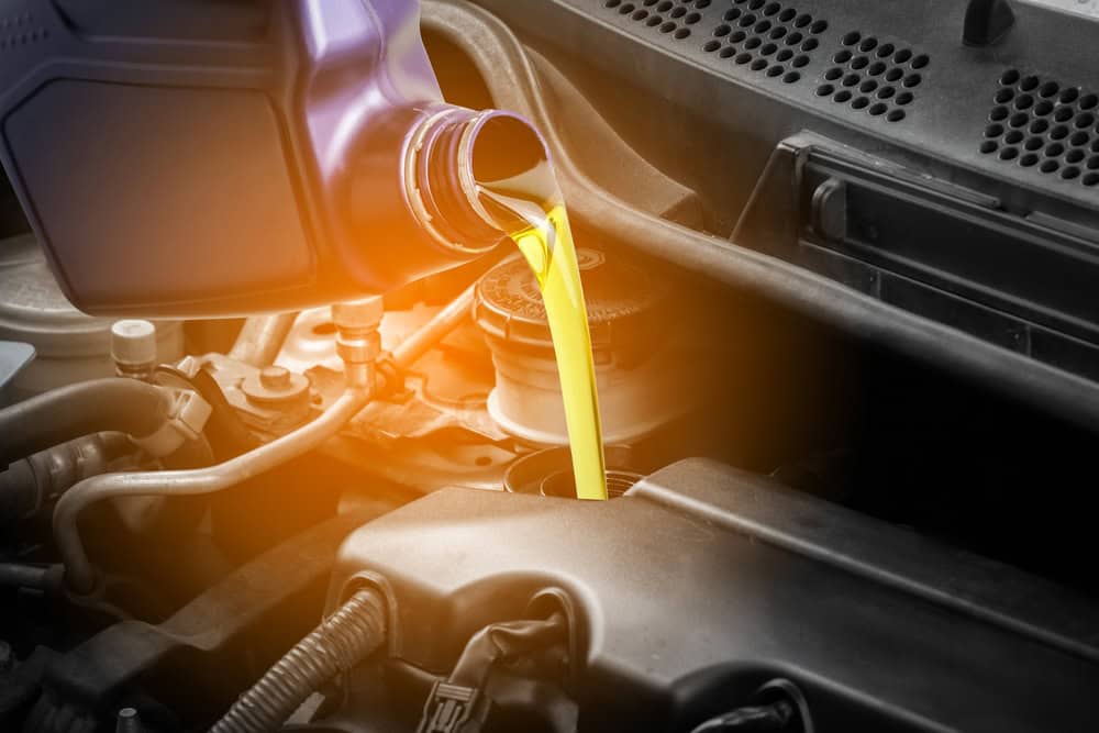 Motor oil pours smoothly from a purple container into the engine compartment of a car, with its bright yellow color highlighted under direct light. Surrounding engine components and hoses are clearly visible, indicating a standard oil change in progress. The setting suggests a professional service environment at Auto Repair Clarke County.