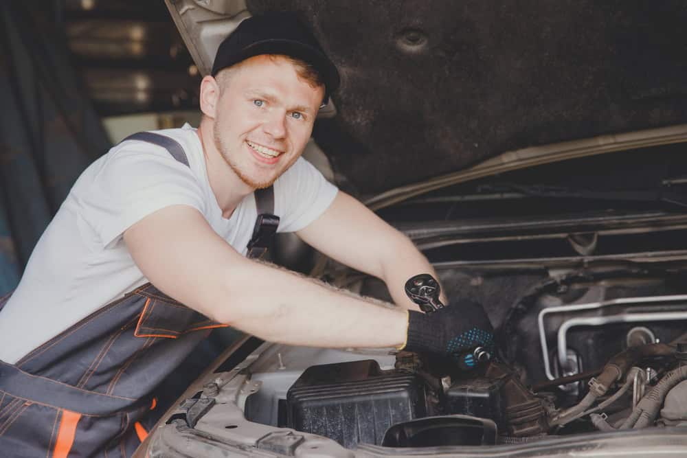 A mechanic in a cap and gloves smiles while working on a car engine. He is wearing a white t-shirt and overalls, with the car hood open. The setting appears to be a garage or workshop.