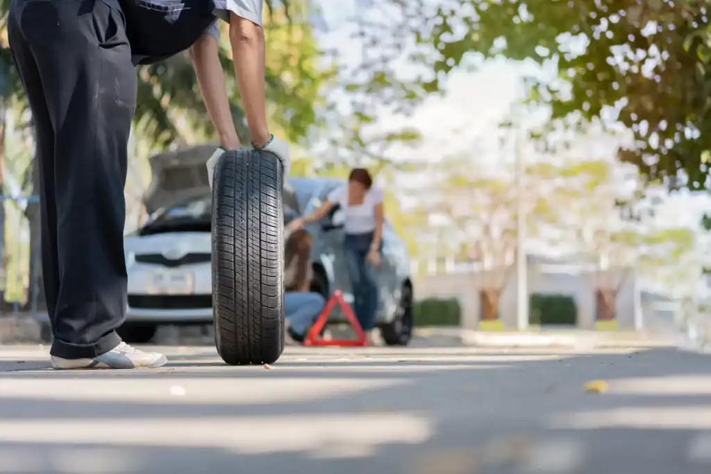 A person rolls a car tire toward another person who is kneeling by a car with the hood open and a warning triangle set up on the road, suggesting a roadside tire change.