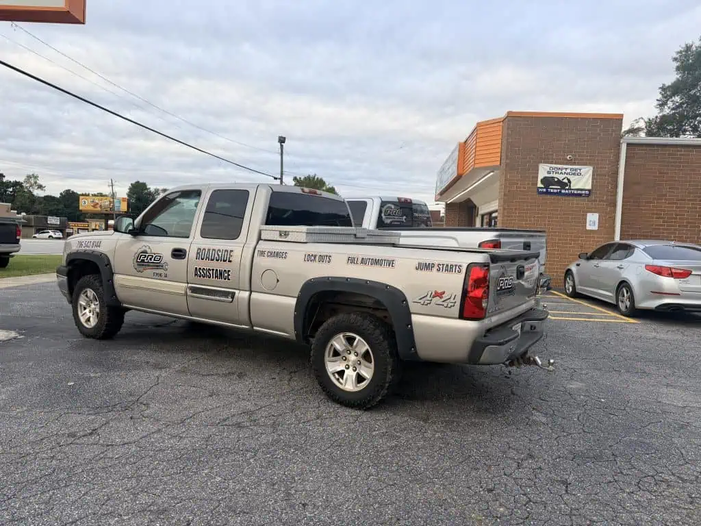 A silver pickup truck with "Roadside Assistance" and "Transmission Rebuilds" written on the side is parked in a lot near a brick building and a Dunkin' store. Another white car is parked nearby.
