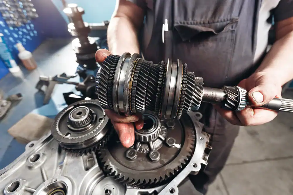 A mechanic wearing a dark uniform holds a set of metal gears, working on disassembling or repairing a vehicle transmission on a workbench with various automotive parts.